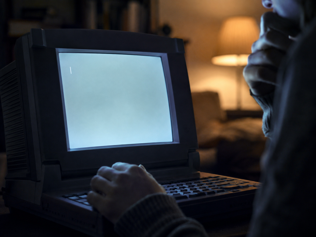 old CRT or early-2000s laptop screen glowing in a dim room, blank text cursor blinking, hands resting near the keyboard mid-thought.